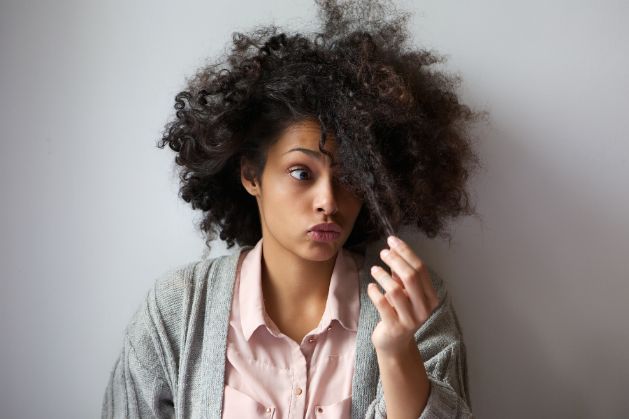 Woman with curly hair touching it, wearing a pink shirt and gray cardigan, against a plain background.
