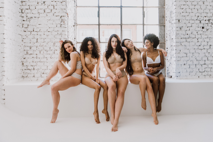 Five women sitting on a white bench against a white brick wall.