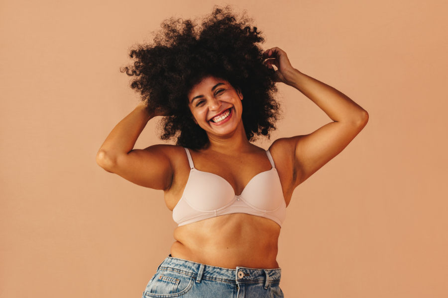 Smiling woman with natural curly afro hair lifting her hair with both hands against a warm beige background, representing confidence and hair wellness.