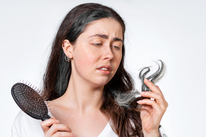 Woman examining a gray strand of hair while holding a hairbrush, illustrating stress and gray hair concerns.